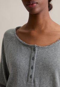 Szara, długorękawowa koszula henley z okrągłym dekoltem, postrzępionymi brzegami oraz czterema ciemnymi guzikami z przodu. Miękka tekstura tkaniny.