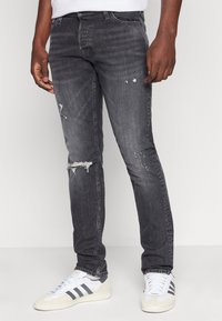 Svarta slitna denimjeans med smal passform, utrustad med revärer vid knäna, subtil blekning och femficksdesign. Matchas med vita sneakers.