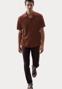 Chemise brune à manches courtes avec un tissu côtelé, assortie à un pantalon slim noir et des sandales noires à brides croisées.