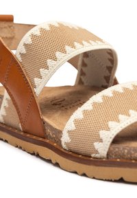 Sandalo in tessuto beige con cinghie incrociate. Presenta dettagli in pelle color nocciola e cuciture a zigzag. Suola in sughero naturale e suola in gomma testurizzata.