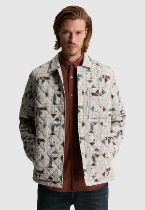 Homme aux cheveux mi-longs portant une veste matelassée avec des motifs géométriques marron et gris sur une chemise bordeaux boutonnée.