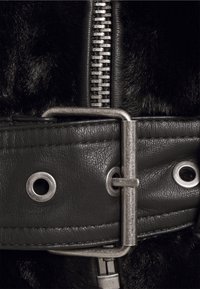 Czarny sztuczny futerko z czarnym skórzanym paskiem z metalową klamrą w kolorze srebrnym i detalami w postaci oczek, obok widoczny srebrny zamek błyskawiczny.