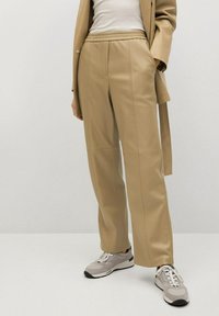 Beige, wijde broek van glad stof, met een trekkoord in de taille en verticale naden, gecombineerd met grijze sneakers.