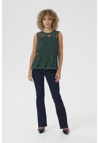 Haut en dentelle verte avec un motif floral, sans manches et à ourlet festonné. Assorti avec un jean évasé bleu foncé et des ballerines noires.