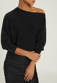 Kobieta w czarnej, błyszczącej bluzce z odkrytymi ramionami i dopasowanej spódnicy, z rękami skrzyżowanymi na talii.