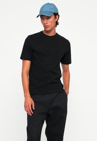 Junger Mann mit blauer Mütze, schlichter schwarzer T-Shirt und schwarzen Hosen, der mit einer Hand in der Tasche vor einem weißen Hintergrund steht.