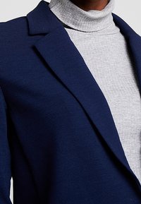 Blazer bleu marine avec revers structurés porté sur un col roulé côtelé gris clair. Le blazer présente un tissu texturé et une coupe ajustée.