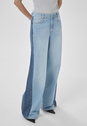 Personne portant un jean à jambes larges avec des panneaux avant clairs et des panneaux latéraux bleu foncé, assorti à des chaussures noires à bouts pointus.