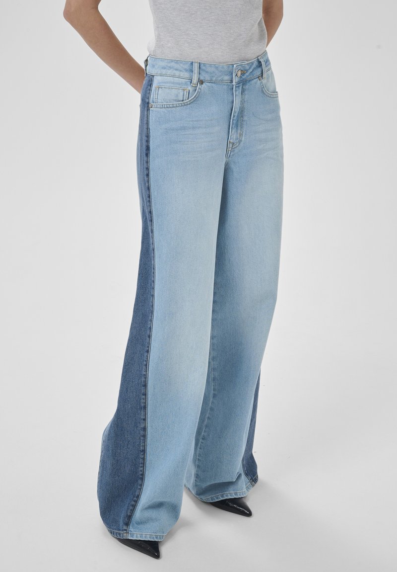 Osoba nosi široké nohavice z džínoviny s ľahkými prednými panelmi a tmavomodrými bočnými panelmi, kombinované s čiernymi topánkami s ostrou špičkou.