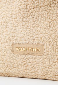 Beige texturerad handväska gjord av fluffigt, mjukt material med en guld "VALENTINO"-logotyp på en slät läderlapp.