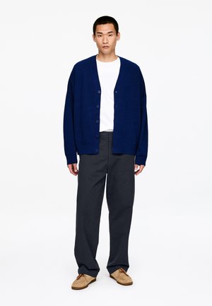 Granatowy kardigan o luźnym kroju, z zapięciem na guziki i ściągaczami na rękawach, zestawiony z ciemnymi spodniami i beżowymi wsuwanymi butami.