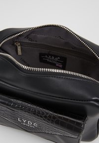 Borsa nera in materiale sintetico con una finitura liscia e un pannello frontale in texture coccodrillo. L'interno presenta una tasca con zip e un'etichetta del marchio. Hardware argentato.