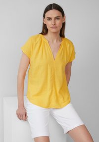 Blouse légère jaune avec un col en V, des manches courtes et des détails froncés au niveau du col. Associée à un short blanc retroussé.