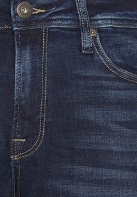 Primo piano di jeans in denim blu scuro che mostrano la tasca anteriore, il bottone, l'anello per la cintura e la cucitura di contrasto gialla.