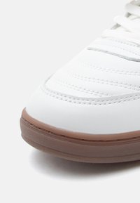 Λευκό αθλητικό παπούτσι με λείο δέρμα στην επάνω πλευρά, ραμμένες λεπτομέρειες και στ�ρογγυλεμένη μύτη. Η σόλα είναι ανοιχτό καφέ και με υφή.