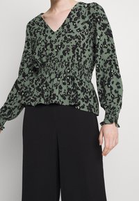 Grüne Bluse mit schwarzem Leopardenmuster, V-Ausschnitt, geraffter Taille und langen Ärmeln mit elastischen Bündchen. Kombiniert mit schwarzen Hosen.