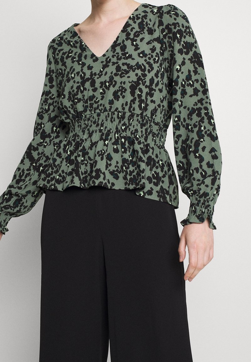 Grüne Bluse mit schwarzem Leopardenmuster, V-Ausschnitt, geraffter Taille und langen Ärmeln mit elastischen Bündchen. Kombiniert mit schwarzen Hosen.
