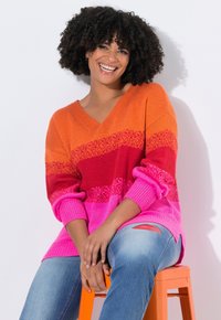 Donna con capelli ricci che sorride, indossando un vivace maglione a strisce arancioni, rosse e rosa e jeans, seduta su uno sgabello arancione contro un muro bianco.