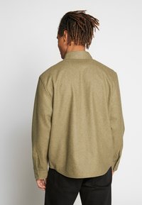 Olijfgroene blouse met lange mouwen en kraag, gemaakt van textuurstof, met knopenmanchetten en een losse pasvorm. Zichtbare stikseldetails op de achterkant.