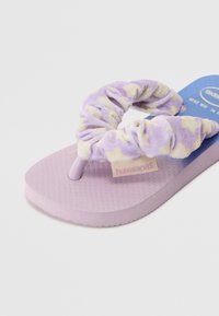 Violetinės šlepetės su tekstūruota pado danga, turinčios minkštą kryžminio modelio dirželį šviesiai violetinės ir geltonos spalvų motyvais, pažymėtos žodžiu 'havaianas'.