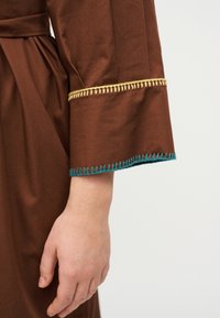 Χέρι ατόμου που φοράει καφέ ένδυμα με κίτρινη και μπλε διακοσμητική ραφή στο φαρδύ μανίκι, σε ομοιόμορφο φόντο.