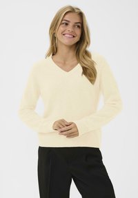 Krämfärgad v-ringad tröja med långa ärmar, ribbade manschetter och fåll, tillverkad av mjukt material, kombinerad med svarta byxor.