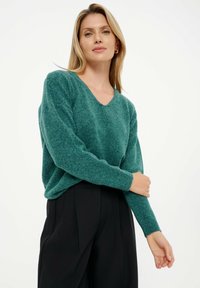 V-hals trui in diep teal, gemaakt van zacht breiwerk. Heeft verlaagde schouders en geribbelde manchetten, gecombineerd met een hoge taille zwarte broek.