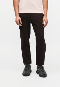 Άτομο που φοράει μαύρο παντελόνι cargo και σκούρα γκρι αθλητικά παπούτσια, όρθιο μπροστά σε απλό λευκό φόντο.