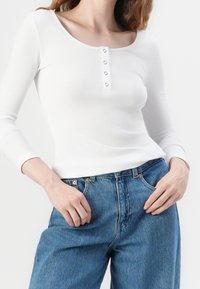 Biała, dług rękawowy, ribbed top z okrągłym dekoltem i pięcioma guzikami. Połączony z niebieskimi dżinsami o luźnym kroju i kieszeniach.