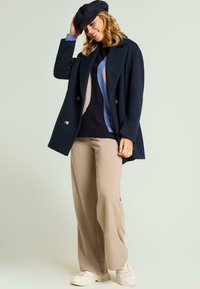 Abrigo de lana azul marino de doble botonadura sobre un suéter de punto con estampado, combinado con pantalones beige de pierna ancha y zapatillas blancas, rematado con una gorra oscura.