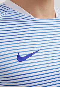 Camisa deportiva a rayas en azul y blanco con cuello redondo y logo de Nike bordado en púrpura en el pecho. Fabricada con material ligero y transpirable.