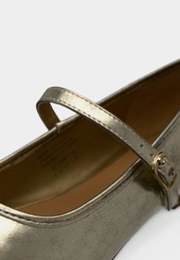 Μία χρυσή μεταλλική γόβα Mary Jane με λεπτό λουράκι και μικρή χρυσή αγκράφα σε απλό λευκό φόντο.