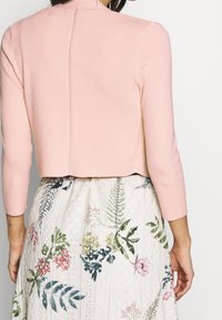 Cardigan corto rosa chiaro con maniche a tre quarti, caratterizzato da una cucitura centrale lungo la schiena. Indossato sopra una gonna bianca con motivi floreali.