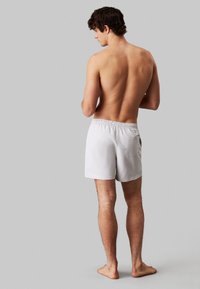 Costumi da bagno grigi chiari da uomo con vita elasticizzata, tasca posteriore e un design semplice. Peluria visibile sulle gambe e piedi nudi.