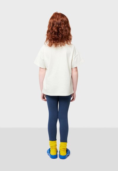 T-shirt corto crema a maniche corte, leggings blu scuro, calzini a coste giallo brillante e zoccoli blu, visti da dietro. Capelli ricci rossi visibili.