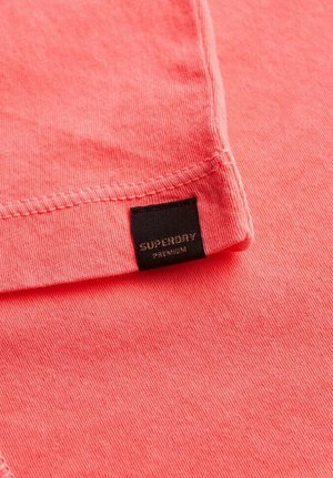Koraalkleurig katoenen T-shirt met een gladde textuur, voorzien van een zwart label met de tekst "SUPERDRY PREMIUM" op de hals.