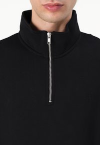 Pulóver negro de media cremallera hecho de tela suave, con un cuello alto, detalle de cremallera y un logotipo bordado en el pecho.
