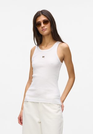 Femme portant des lunettes ovales marron, un haut blanc sans manches avec un petit motif sur la poitrine et un pantalon blanc, debout devant un fond uni.