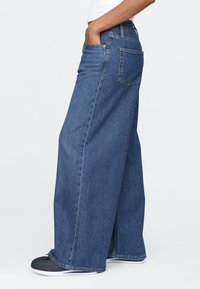 Traperice širokih nogavica u srednje plavoj boji, s visokim strukom, bočnim džepovima i teksturiranim završetkom. Ukrašene standardnim džepom straga.