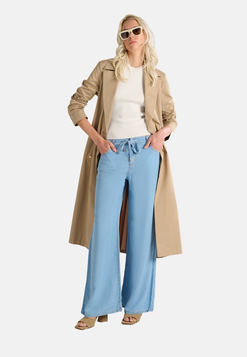 Trench coat lungo beige, top bianco aderente, jeans ampi blu chiaro con coulisse e sandali con tacco beige. Gli occhiali da sole completano il look.