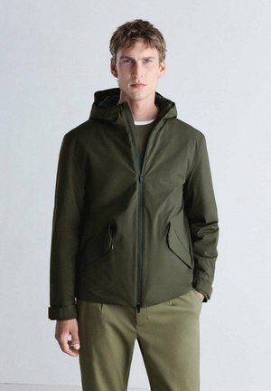 Man with short hair wearing a dark green zip-up jacket with a hood and olive-green pants, standing against a plain light background.