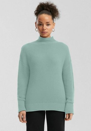 Frau mit lockigem, hochgebundenem Haar, die Creolen trägt und einen hellblauen, gestrickten Rollkragenpullover sowie schwarze Hose anhat.