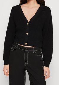 Zwarte cropped cardigan met een diepe V-hals, geribbelde textuur en drie goudkleurige knopen. Gecombineerd met hooggetailleerde zwarte broek met witte stikselaccenten.