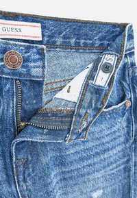 Jean en denim avec un délavage bleu clair, présentant une fermeture éclair, des coutures contrastées et des détails d'usure visibles près des poches et de la taille.