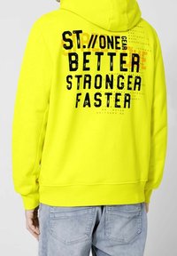 Felpa con cappuccio di un giallo acceso, con un audace testo nero sul retro, polsini e vita a costine. Il materiale sembra morbido e strutturato.