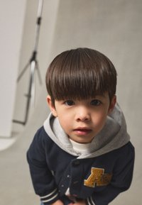 Bambino con una giacca varsity blu navy con cappuccio grigio, con una lettera 'A' dorata ricamata. Capelli corti, lisci e scuri, sfondo neutro.