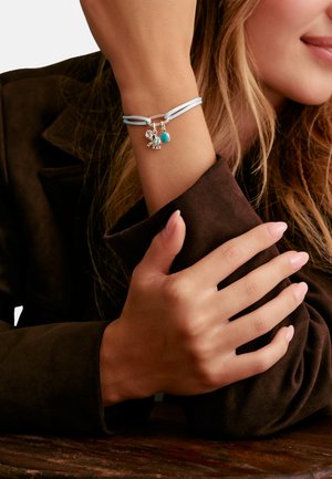 Frau trägt ein hellblaues Cord-Armband mit silbernen Elefanten- und Türkisstein-Anhängern, legt ihren Arm auf eine Holzoberfläche und trägt eine braune Jacke.