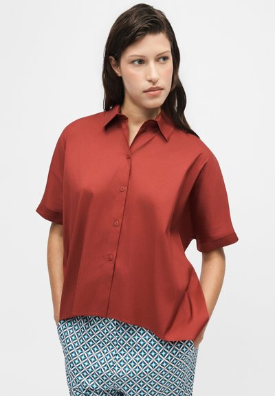 Femme aux cheveux foncés portant une chemise rouge ample à manches courtes boutonnée et un pantalon bleu à motifs, regardant sur le côté, les mains dans les poches.