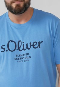 Lichtblauw katoenen T-shirt met een ronde hals en een donkerblauw bedrukt logo. De tekst luidt "s.Oliver Elevated Essentials Since 1969."