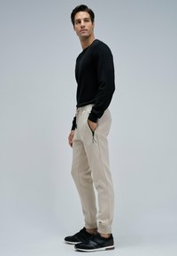 Hombre de pie con el torso ligeramente de lado, viste una camiseta de manga larga negra, pantalones jogging beige con bolsillos con cremallera y zapatillas negras, todo sobre un fondo gris claro.